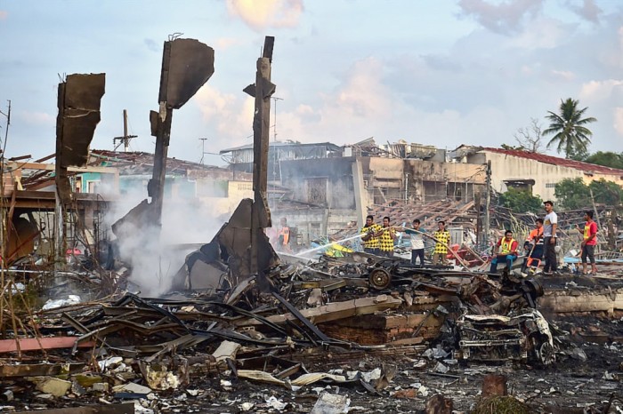 Petugas pemadam kebakaran Thailand memadamkan bara api di sekitar rumah yang hancur setelah ledakan merobek gudang kembang api, menewaskan duabelas orang dan melukai lebih dari 121 orang, di distrik Sungai Kolok di provinsi Narathiwat, Thailand selatan Sabtu (29/7/2023). (Foto: AFP/Madaree T)